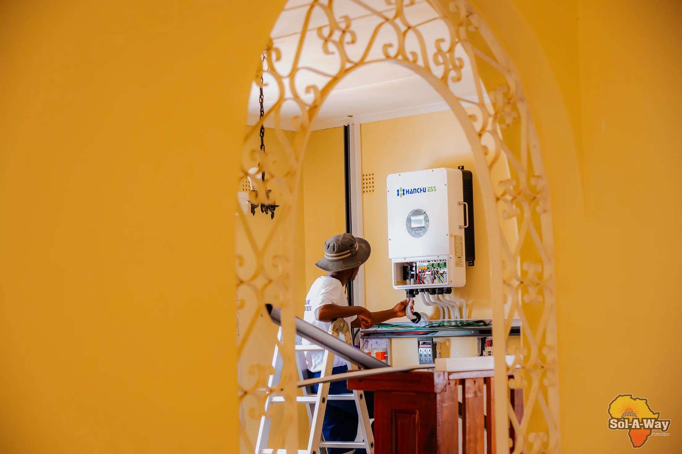 Technician installing a hybrid inverter on a yellow wall, seen through an archway